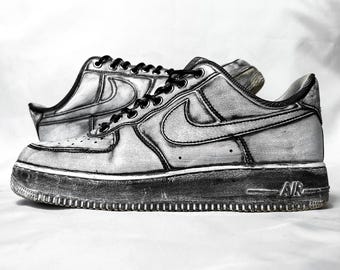 May include: A pair of low-top sneakers featuring a monochromatic, hand-drawn aesthetic with bold black outlines on a light gray base. The shoes include black laces, a prominent side swoosh, and the word AIR embossed on the textured rubber sole.