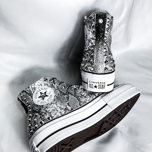 Può includere: Scarpe da ginnastica alte Converse All Star con motivo a pelle di serpente e borchie argentate. Le scarpe hanno una suola bianca e una suola in gomma marrone. Il logo Converse All Star è visibile.