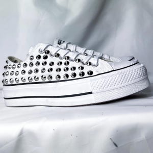 Puede incluir: Zapatillas Converse blancas de plataforma con tachuelas plateadas que cubren toda la parte superior. Las zapatillas tienen un borde negro alrededor de la suela.