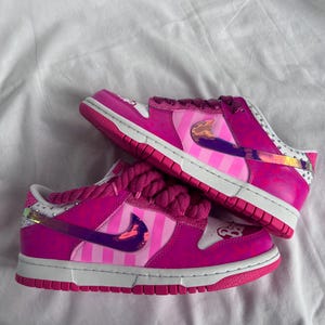 May include: Pair of pink and white sneakers with a striped design on the side panels. The shoes feature a pink sole, laces, and accents, with a white midsole and a decorative element on the tongue.