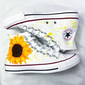 Puede incluir: Zapatillas Converse blancas de caña alta con un girasol amarillo pintado en el lateral. Las zapatillas tienen salpicaduras de pintura amarilla y pedrería en el lateral. El logotipo de Converse es visible en el lateral de la zapatilla.