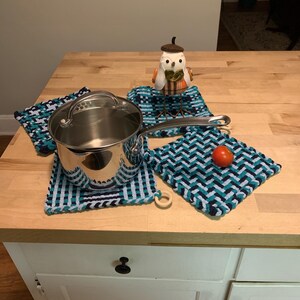 May include: A stainless steel pot with a glass lid sits on a blue and white woven pot holder.  A second blue and white woven pot holder is next to the pot, and a third blue and white woven pot holder is in the background.  A small ceramic owl figurine is on top of the pot holder in the background.