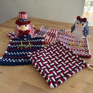 May include: Three woven pot holders in red, white, and blue with a patriotic theme. Two of the pot holders feature a woven checkered pattern, and the third pot holder features a woven herringbone pattern. Two handmade bird figurines are on top of the pot holders. One bird is wearing a red, white, and blue hat, and the other bird is wearing a red bow.