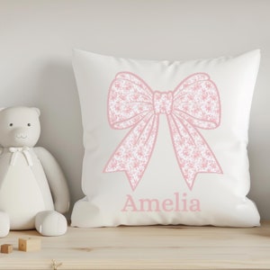 May include: White square pillow with a large pink bow design and the name "Amelia" printed in pink. The bow has a floral pattern. The pillow is on a wooden shelf with stuffed animals.