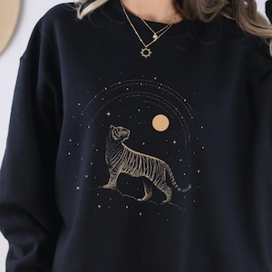 Op de afbeelding: Zwart sweatshirt met een gouden tijgerontwerp en hemelse elementen. De tijger is gedetailleerd, tegen een achtergrond van sterren en een maan. Het woord "BLACK" staat onderaan gedrukt. Een casual, comfortabel kledingstuk.