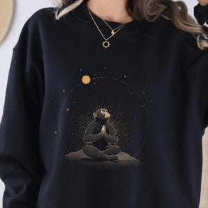 May include: Black sweatshirt featuring a graphic of a meditating monkey with a celestial design. The design includes a sun and stars. The word "BLACK" is printed at the bottom left of the image.