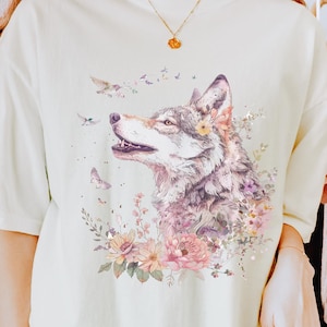 Pode incluir: Uma camiseta branca com um desenho de aquarela de um lobo cercado por flores. O lobo está olhando para a esquerda e tem uma coroa de flores na cabeça. O texto "Marfim" é visível no canto inferior direito da imagem.
