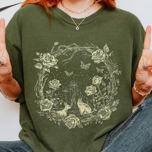 May include: Olive green sweatshirt with a circular floral design featuring roses, butterflies, a deer, and a cat. The word "HEMP" is printed in white on the upper right. The model is wearing blue jeans and white sneakers.