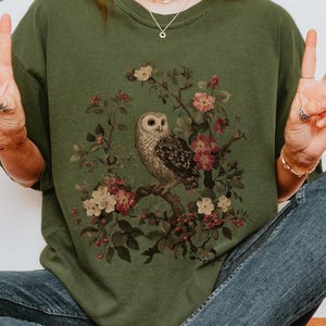 May include: An olive green sweatshirt featuring a detailed illustration of an owl perched on a flowering branch. The owl is depicted with intricate feather patterns, surrounded by pink and cream-colored blossoms. The word "HEMP" is printed in white.