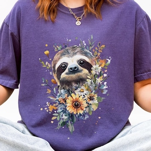 Peut inclure: Un t-shirt violet avec une illustration à l'aquarelle d'un paresseux entouré de fleurs. Le paresseux a une fourrure brune et une expression amicale. Les fleurs comprennent des tournesols et d'autres fleurs colorées. Le mot "GRAPE" est imprimé en haut à droite.