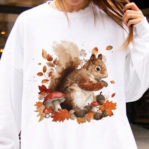 May include: White sweatshirt with a watercolour illustration of a squirrel amidst autumn leaves, acorns, and mushrooms. The squirrel is brown and tan, with a fluffy tail. The design is centered on the front. The word "WHITE" is printed at the bottom right.