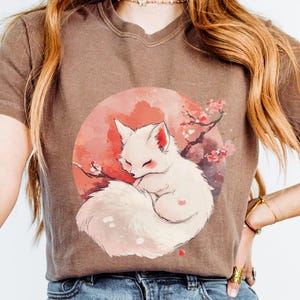 Peut inclure: Un t-shirt marron avec un renard blanc enroulé dans un cercle de fleurs roses et orange. Le renard est entouré d'un cercle rose et orange avec un contour blanc. Le t-shirt a un col rond et des manches courtes.