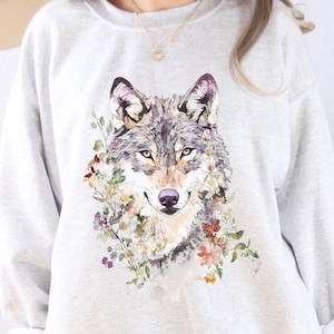 Pode incluir: Camisola cinza claro com uma ilustração em aquarela detalhada da cabeça de um lobo rodeada por flores coloridas. A palavra "ASH" está impressa em preto. A camisola tem gola redonda e mangas compridas.