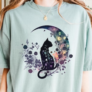 May include: A light green t-shirt featuring a black cat silhouette sitting inside a crescent moon. The moon is filled with colorful floral and botanical elements. The shirt has a relaxed fit.