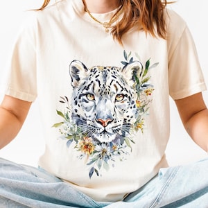 May include: Ivory t-shirt with a watercolour illustration of a snow leopard's face and floral elements. The word "IVORY" is in the top right corner. The model wears light blue jeans and white trainers.