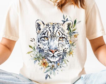 Könnte beinhalten: Elfenbeinfarbenes T-Shirt mit einer Aquarellillustration eines Schneeleoparden-Gesichts und floralen Elementen. Das Wort "IVORY" steht oben rechts. Das Model trägt hellblaue Jeans und weiße Turnschuhe.