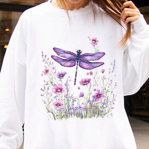Può includere: Felpa bianca con un design di libellula viola e fiori. La grafica mostra una libellula con le ali spiegate, circondata da fiori viola e rosa e fogliame verde. La parola "WHITE" è stampata in basso a destra.