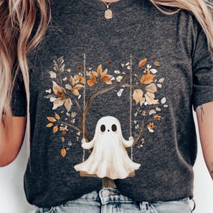 Könnte beinhalten: Dunkelgraues meliertes T-Shirt mit einem Geist auf einer Schaukel vor einem Hintergrund aus Herbstblättern und -zweigen. Der Text "DARK GRAY HEATHER" ist auf der linken Seite des Shirts aufgedruckt.