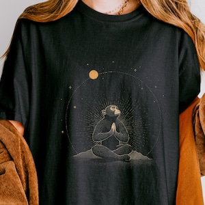 May include: Black t-shirt featuring a golden illustration of a meditating monkey within a celestial design. The monkey is seated with hands clasped in prayer. The word "BLACK" is printed in white on the upper right. The shirt is a comfortable, casual style.