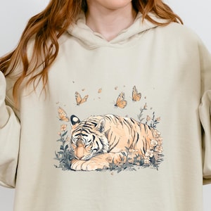 Può includere: Felpa con cappuccio color sabbia con un'illustrazione ad acquerello di una tigre che riposa tra fiori e farfalle. Il design include una tigre a strisce arancioni e nere, circondata da elementi floreali e farfalle. La parola "SAND" è stampata in basso.