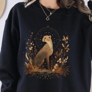 May include: Black sweatshirt featuring a detailed illustration of a cheetah surrounded by golden foliage and a starry circle. The cheetah is depicted in a seated position. The word "BLACK" is printed at the bottom of the sweatshirt.