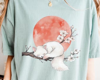 May include: A pale blue t-shirt with a graphic of a sleeping white fox on a branch with cherry blossoms and a red sun. The word "BAY" is printed in white on the right side of the shirt.