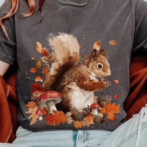 May include: Dark gray t-shirt featuring a detailed illustration of a squirrel surrounded by autumn leaves, acorns, and red-capped mushrooms. The word "PEPPER" is printed at the top right. The shirt has short sleeves.
