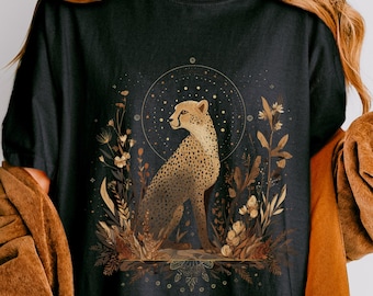 以下が含まれることがあります： 黒いTシャツ。チーターのイラストが描かれています。チーターは座った姿勢で描かれ、花の要素と天体の背景に囲まれています。「BLACK」の文字がTシャツの右上部に白で印刷されています。