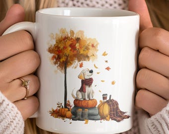 May include: White ceramic mug featuring an autumn-themed illustration. The design includes a tree with orange and yellow leaves, a dog wearing a scarf, pumpkins, and a plaid blanket. The mug is held by a person wearing a cream-colored sweater.