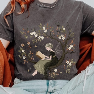 May include: Dark gray t-shirt featuring a whimsical illustration of a fairy reading a book while perched on a flowering tree branch. The word "PEPPER" is printed at the top right. The fairy is wearing a black dress and has delicate wings.