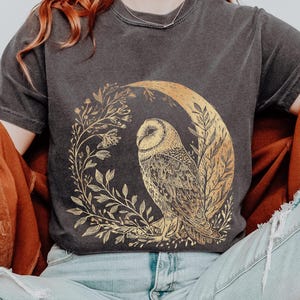 May include: Dark gray t-shirt featuring a gold illustration of an owl perched on a crescent moon, surrounded by floral elements. The word "PEPPER" is printed at the top right. The shirt has short sleeves and a crew neck.