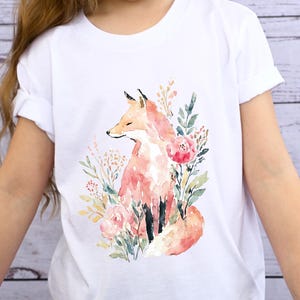 May include: White t-shirt featuring a watercolor illustration of a fox surrounded by flowers and foliage. The fox is depicted in shades of orange and pink, with green and yellow accents in the floral arrangement. The word "WHITE" is printed on the shirt.