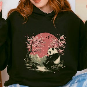 May include: Black hoodie featuring a panda illustration in a serene landscape. The design includes a panda sitting under a cherry blossom tree with a pink sun in the background. The hoodie is made of a soft material.