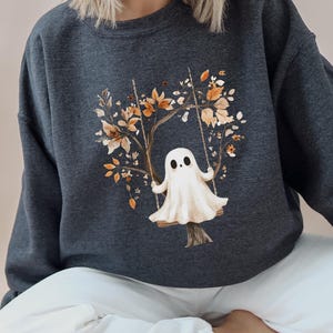 May include: Dark heather gray sweatshirt featuring a watercolor illustration of a ghost on a swing. The swing is attached to a tree with autumn leaves in shades of orange and brown. The text "DARK HEATHER" is printed on the left side of the sweatshirt.