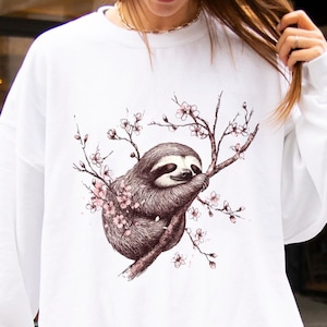May include: White sweatshirt featuring a brown sloth perched on a flowering branch. The sloth is surrounded by pink blossoms. The word "WHITE" is printed in white at the bottom right of the image.