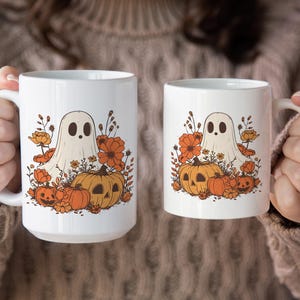 May include: Two white ceramic mugs with a Halloween-themed design. Each mug features a cartoon ghost, pumpkins, and orange flowers. The mugs are held by a person wearing a brown sweater.
