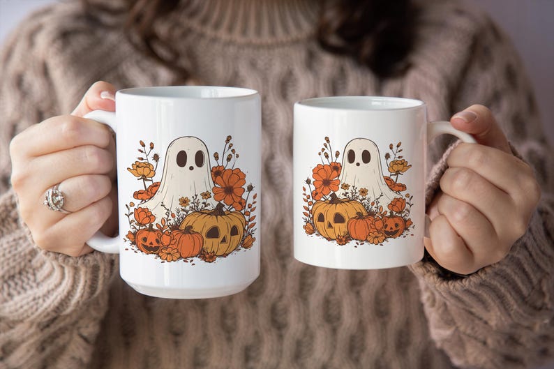 May include: Two white ceramic mugs with a Halloween-themed design. Each mug features a cartoon ghost, pumpkins, and orange flowers. The mugs are held by a person wearing a brown sweater.