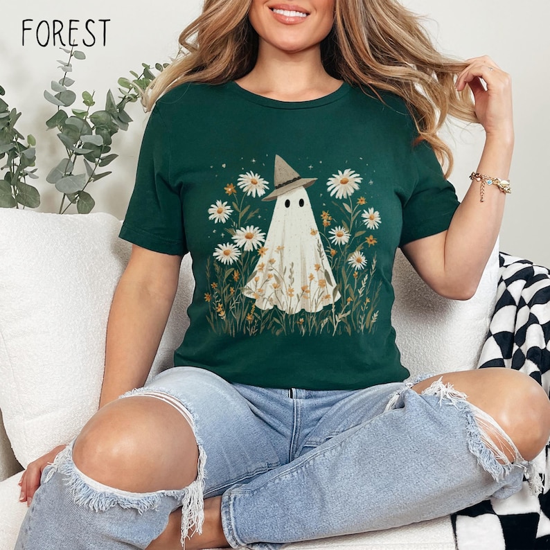 K&ouml;nnte beinhalten: Dunkelgr&uuml;nes T-Shirt mit einem skurrilen Geisterdesign, das einen Hexenhut tr&auml;gt, umgeben von G&auml;nsebl&uuml;mchen und Wildblumen. Das Wort "FOREST" ist oben links aufgedruckt. Das Shirt wird mit hellblauen Jeans kombiniert.