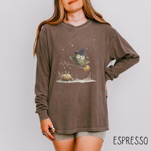 May include: Espresso-colored long-sleeve shirt featuring a whimsical owl wearing a witch hat and holding a pumpkin. The graphic includes a pumpkin and stars. The word "ESPRESSO" is printed at the bottom of the shirt.