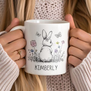 May include: White ceramic mug with a black and white illustration of a rabbit in a field of flowers. The mug has the name "KIMBERLY" printed on the bottom.