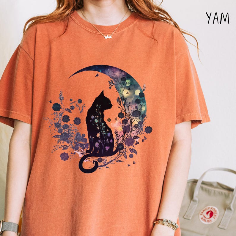 May include: Orange t-shirt featuring a black cat silhouette sitting inside a crescent moon with floral and celestial details. The word "YAM" is printed on the upper right side of the shirt. The shirt has short sleeves and a relaxed fit.