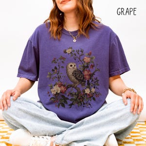 May include: A purple t-shirt with a vintage-style owl and floral design. The owl is perched on a branch surrounded by pink and white flowers. The word "GRAPE" is printed in black in the upper right corner.