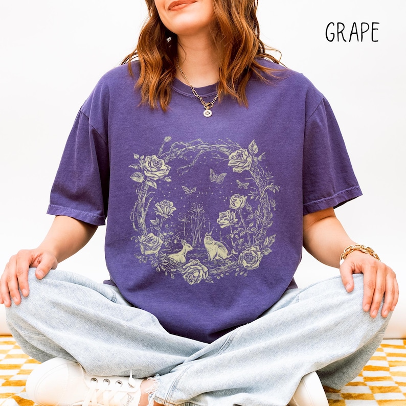 May include: A purple t-shirt with a floral and animal graphic. The design features roses, butterflies, a deer, and a cat within a circular wreath. The word "GRAPE" is printed in the upper right corner.
