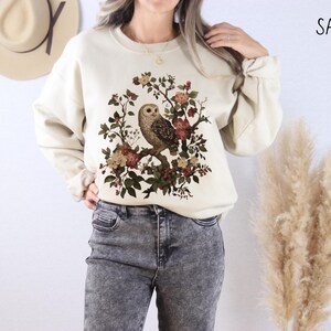 Op de afbeelding: Zandkleurige sweatshirt met een gedetailleerde uilillustratie die op een bloemen tak zit. Het ontwerp bevat een verscheidenheid aan bloemen in tinten rood, bruin en cr&egrave;me. Het woord "SAND" is in het zwart rechtsboven gedrukt.
