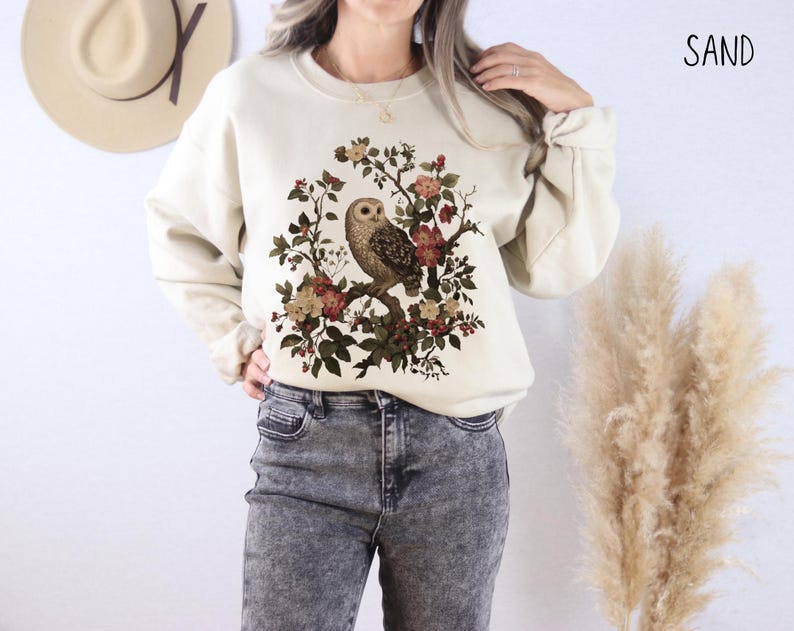 Op de afbeelding: Zandkleurige sweatshirt met een gedetailleerde uilillustratie die op een bloemen tak zit. Het ontwerp bevat een verscheidenheid aan bloemen in tinten rood, bruin en cr&egrave;me. Het woord "SAND" is in het zwart rechtsboven gedrukt.