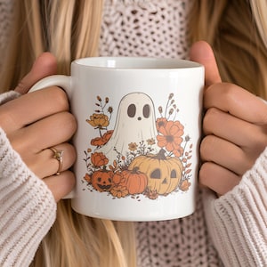May include: White ceramic mug featuring a whimsical Halloween design. The design includes a friendly ghost, pumpkins, and orange flowers. The mug is held by a person wearing a cream-colored sweater.