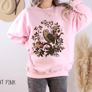 Op de afbeelding: Lichtroze sweatshirt met een gedetailleerde uil illustratie op een bloeiende tak. De uil is omringd door bloemen in rood, geel en groen. De tekst "LIGHT PINK" staat onderaan.