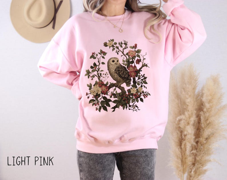 Op de afbeelding: Lichtroze sweatshirt met een gedetailleerde uil illustratie op een bloeiende tak. De uil is omringd door bloemen in rood, geel en groen. De tekst "LIGHT PINK" staat onderaan.