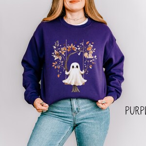 May include: A purple sweatshirt featuring a whimsical illustration of a ghost on a swing set, surrounded by autumn leaves. The word "PURPLE" is printed in black on the lower right side. The sweatshirt is made of a soft material.