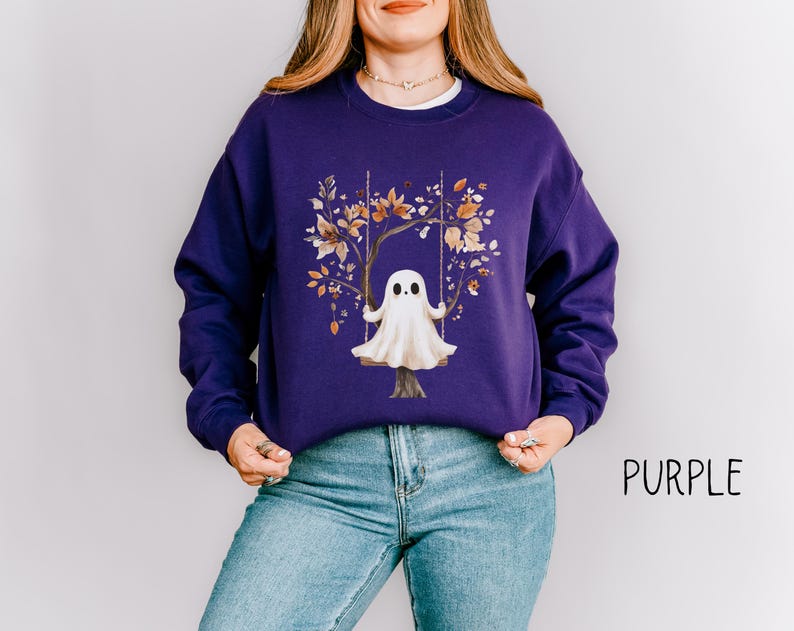 May include: A purple sweatshirt featuring a whimsical illustration of a ghost on a swing set, surrounded by autumn leaves. The word "PURPLE" is printed in black on the lower right side. The sweatshirt is made of a soft material.